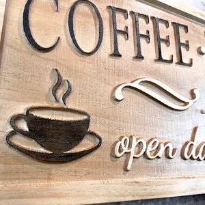 K&ouml;nnte beinhalten: Ein Holzschild mit dem Text "Coffee - Tea - Bakery" und "open daily" in einer dekorativen Schriftart. Ein stilisiertes Bild einer Kaffeetasse mit aufsteigendem Dampf befindet sich in der Mitte des Schildes.