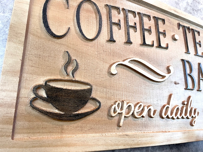 K&ouml;nnte beinhalten: Ein Holzschild mit dem Text "Coffee - Tea - Bakery" und "open daily" in einer dekorativen Schriftart. Ein stilisiertes Bild einer Kaffeetasse mit aufsteigendem Dampf befindet sich in der Mitte des Schildes.