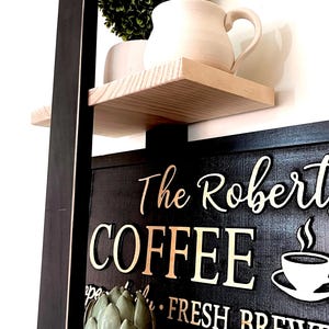 May include: A black wooden sign with white lettering that reads "The Robert's Coffee Fresh Brew". The sign is on a shelf with a small green plant and a white ceramic mug.