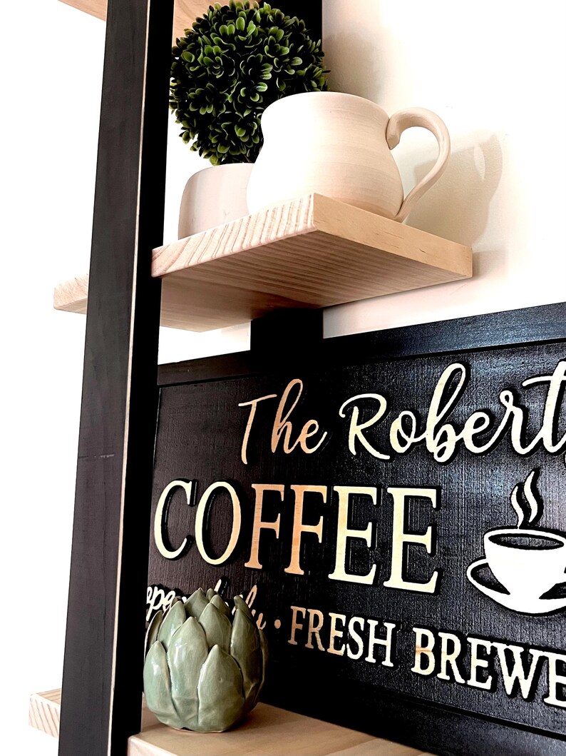 May include: A black wooden sign with white lettering that reads "The Robert's Coffee Fresh Brew". The sign is on a shelf with a small green plant and a white ceramic mug.