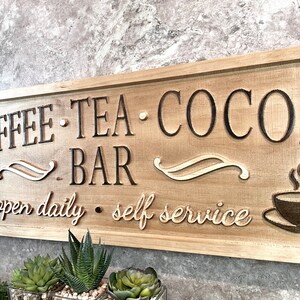 K&ouml;nnte beinhalten: Ein Holzschild mit dem Text "COFFEE TEA COCOA BAR open daily self service" in schwarzer Schrift. Das Schild hat eine braune Holzmaserung und zeigt zwei stilisierte Kaffeetassen mit aufsteigendem Dampf.