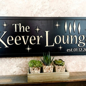 May include: Black wooden sign with gold lettering that reads "The Keever Lounge est. 01.12.2021". The sign is decorated with gold stars and diamonds. Three small potted succulents are in front of the sign.