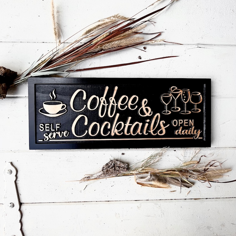 May include: Black wooden sign with gold lettering that reads "Coffee & Cocktails" with a coffee cup and steam icon on the left and a row of four glasses on the right. The sign also says "Self Serve" and "Open Daily".