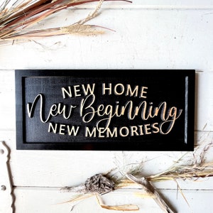 May include: Black wooden sign with gold lettering that reads "New Home New Beginning New Memories".