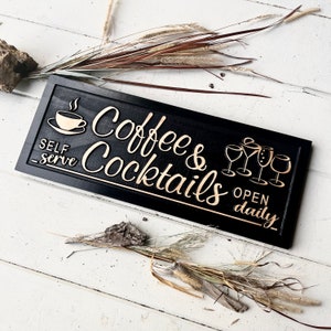 May include: Black wooden sign with gold lettering that reads "Coffee & Cocktails" and "Open Daily". The sign also features a gold coffee cup and a set of gold wine glasses.