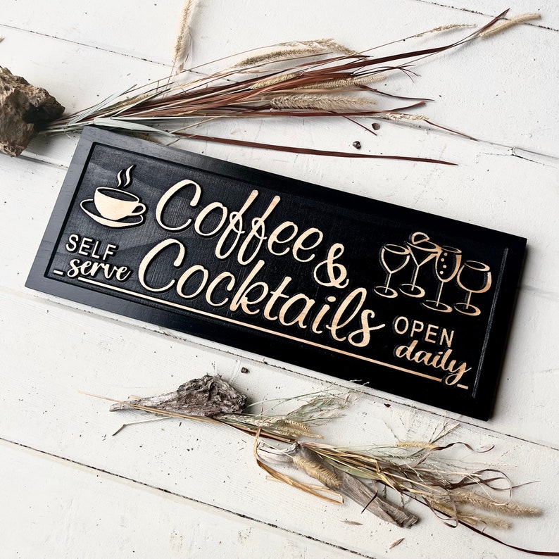 May include: Black wooden sign with gold lettering that reads "Coffee & Cocktails" and "Open Daily". The sign also features a gold coffee cup and a set of gold wine glasses.