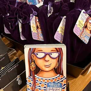 May include: Purple velvet pouches with the text "The Geeky Girl Oracle Deck" printed on a tag. The deck features a cartoon illustration of a girl with glasses and a colorful shirt. The text "Hello, my name is Geeky Girl" is printed on a name tag.