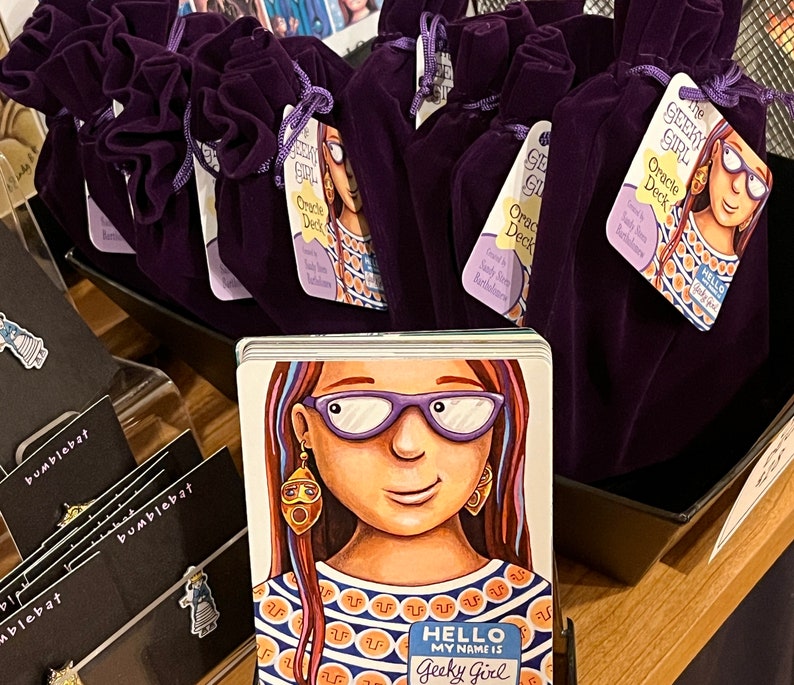 May include: Purple velvet pouches with the text "The Geeky Girl Oracle Deck" printed on a tag. The deck features a cartoon illustration of a girl with glasses and a colorful shirt. The text "Hello, my name is Geeky Girl" is printed on a name tag.