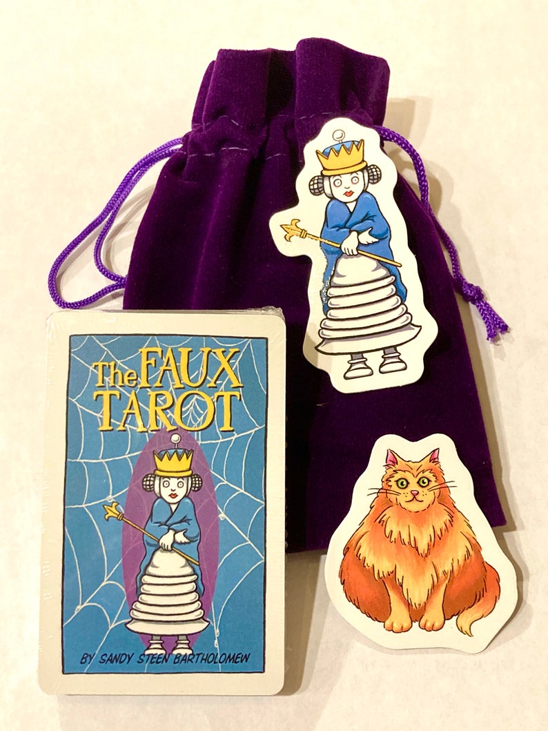 May include: A purple velvet bag with a drawstring closure contains a deck of cards called "The Faux Tarot" by Sandy Steen Bartholomew. The cards feature a cartoon illustration of a queen with a crown and a scepter. Two stickers are also included, one with a cartoon cat and the other with a cartoon queen.