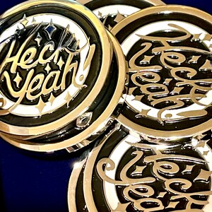May include: Three silver-colored round metal pins with a black and white design. The pins feature the text "Heck Yeah!" in a stylized font.