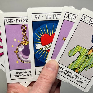 May include: Four tarot cards with purple backgrounds and colorful illustrations. The cards are titled "XV. The Splat", "XXIX. The Crystal", "XV. The Tattoo", and "X. The Scarecrow". The cards feature a pink splatter, a crystal ball on a chain, a heart with the word "Forever" on it, and a scarecrow with a crow on its head.