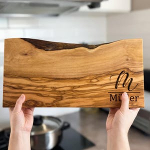Può includere: Tagliere rettangolare in legno chiaro con bordo naturale. Il nome "Müller" è inciso in corsivo. Il tagliere è tenuto in mano da due mani.