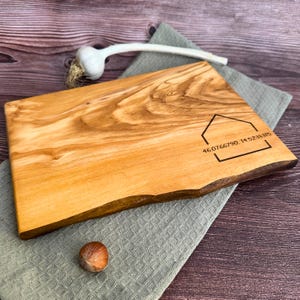 May include: A rectangular wooden chopping board with a natural edge. The board is light brown with darker wood grain patterns. A house outline with coordinates is etched on the board. A bulb of garlic and a hazelnut are also present.