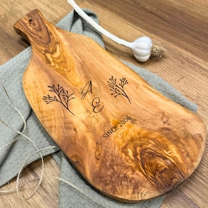 Könnte beinhalten: Ein Holzschneidebrett mit natürlicher Maserung. Das Brett hat einen Griff und ist mit den Initialen "A E", floralen Designs und dem Text "SINCE 2006" graviert. Eine Knoblauchzehe und ein graues Tuch befinden sich im Hintergrund.