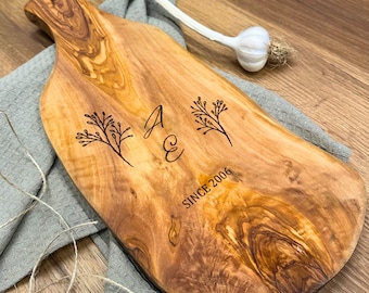 May include: A wooden cutting board with a natural wood grain pattern. The board has a handle and is engraved with the initials "A E", floral designs, and the text "SINCE 2006". A bulb of garlic and a gray cloth are in the background.