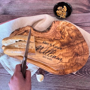Puede incluir: Una tabla de cortar de madera natural grande con una forma única y un patrón de veta de madera natural. La tabla está grabada con el texto "Alex & Laura William -EST 2020-". Se está cortando una barra de pan en la tabla con un cuchillo.