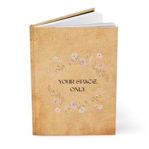 Könnte beinhalten: Ein beigefarbenes Notizbuch mit strukturiertem Einband, verziert mit einem floralen Kranz mit rosa und weißen Blüten. Der Text "YOUR SPACE. ONLY." ist im Kranz zentriert. Das Notizbuch ist geöffnet und zeigt weiße Seiten.