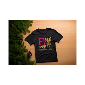 Puede incluir: Camiseta negra con un gráfico rosa y dorado. El gráfico incluye las letras "F7", letras griegas y un diseño de fénix. El texto "Happy Founder's Day" está debajo. La camiseta se muestra sobre una superficie de madera.