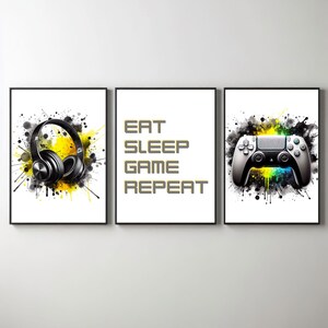Może przedstawiać: Trzy oprawione wydruki z motywem gier. Lewy wydruk przedstawia czarne słuchawki z żółtymi plamami farby. Środkowy wydruk zawiera tekst "EAT SLEEP GAME REPEAT" żółtymi literami. Prawy wydruk przedstawia biały kontroler do gier z żółtymi i niebieskimi plamami farby.