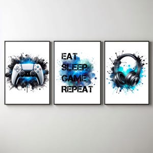 May include: Three black framed prints with blue watercolor splashes. The left print features a white video game controller. The middle print has the text "EAT SLEEP GAME REPEAT". The right print features black headphones.