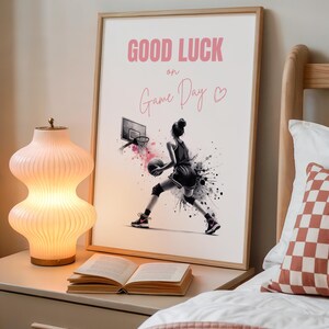 Może przedstawiać: Oprawiony wydruk z różowym napisem "GOOD LUCK on Game Day". Grafika przedstawia koszykarkę w akcji, z efektem akwareli w czerni i bieli. Na stoliku nocnym stoi zapalona, różowa lampa z żebrami i otwarta książka.