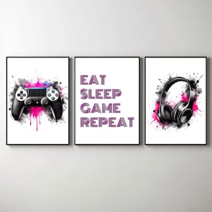 May include: Three framed prints with a gamer theme. The left print features a black and white video game controller with pink watercolor splatters. The middle print has the text "EAT SLEEP GAME REPEAT" in purple. The right print shows a pair of black headphones with pink watercolor splatters.