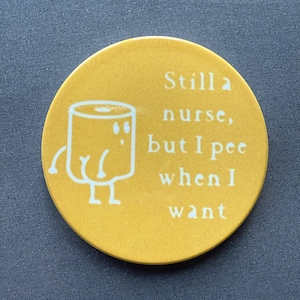 Puede incluir: Posavasos redondo amarillo con un dibujo animado blanco de un inodoro y el texto "Todavía una enfermera, pero meo cuando quiero."