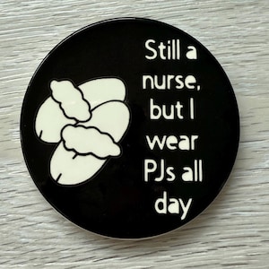 以下が含まれることがあります： 「Still a nurse, but I wear PJs all day」というテキストが書かれた黒と白の丸いコースターです。