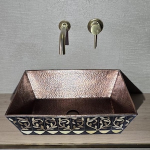 Könnte beinhalten: Ein rechteckiges Waschbecken aus Kupfer mit gehammerter Textur und dekorativem floralen Muster auf der Vorderseite. Das Waschbecken ist unter zwei Wasserhähnen aus gebürstetem Nickel montiert. Ein einzigartiges Badezimmer-Element.