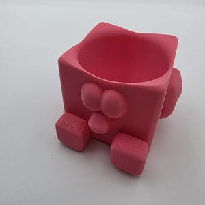 Puede incluir: Una maceta rosa en forma de cubo con elementos redondeados, incluyendo orejas, nariz y pies. La maceta tiene una abertura circular en la parte superior para las plantas. El diseño es caprichoso y juguetón.