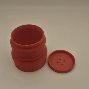 Puede incluir: Un recipiente cilíndrico rojo con una tapa a juego. El recipiente tiene un diseño texturizado y acanalado y la tapa se asemeja a un botón grande con cuatro agujeros. Los artículos están hechos de un material sólido y opaco.