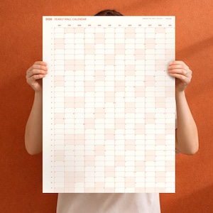 May include: A large, rectangular 2026 yearly wall calendar with a grid of squares in shades of peach and white. The calendar is held up by a person against an orange background. The text "2026 YEARLY WALL CALENDAR" is at the top.