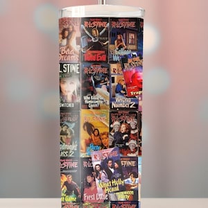 Puede incluir: Un vaso de acero inoxidable con un collage de portadas de libros de "Pesadillas" de R.L. Stine. El vaso tiene una pajita plateada y una tapa blanca. Las portadas son de varios colores y presentan títulos como "Bad Dreams" y "What Holly Heard".