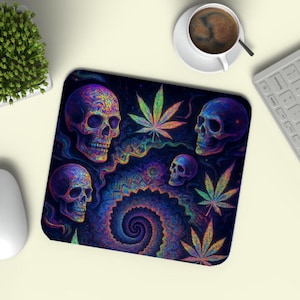 May include: A psychedelic mousepad featuring a dark background with colorful skulls, cannabis leaves, and a spiral design. The mousepad is surrounded by a white mouse, keyboard, a cup of coffee, and a small plant.