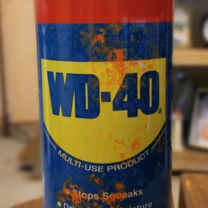 Może przedstawiać: Cylindryczny kubek z czerwoną górą i niebieską podstawą, z logo WD-40 i informacjami o produkcie. Projekt zawiera tekst o wielofunkcyjnych możliwościach produktu. Kubek ma pojemność około 100 ml.