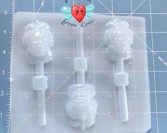May include: Clear plastic lollipop mold with three skull-shaped cavities. Each cavity has a stick and a rectangular support. The mold is on a blue grid surface. The text "Molded from Love" is in the center.