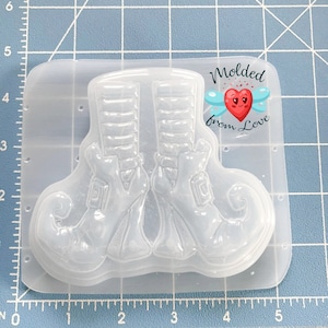 May include: Clear plastic mold of a pair of elf shoes with pointed toes. The mold is on a blue grid background. The text "Molded from Love" is on the mold.