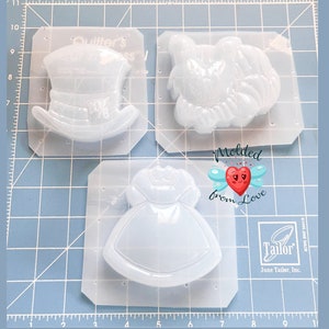 May include: A clear plastic mold with three cavities for making resin crafts. The cavities are shaped like a hat, a dress, and a bird. The mold is labeled "Quilter's Choice" and "Molded from Love".