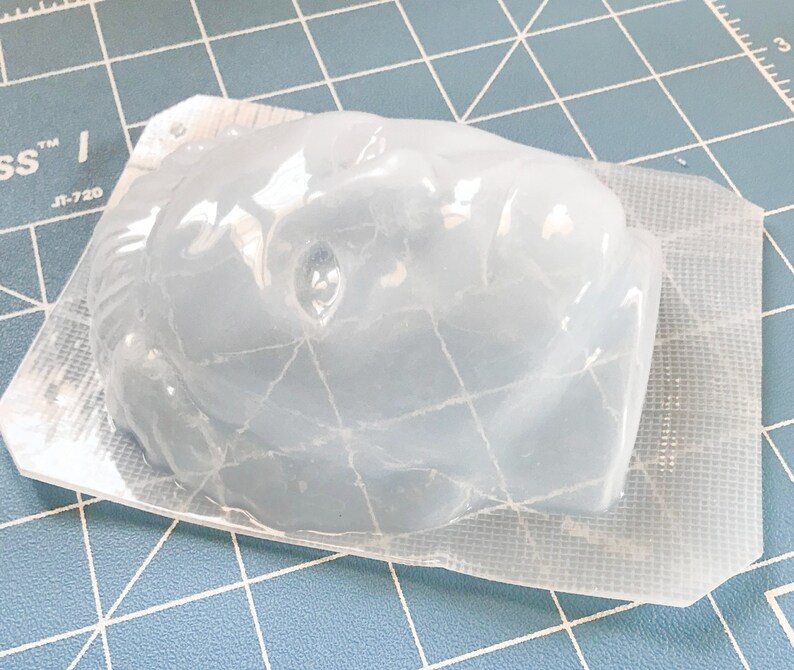 May include: A clear, molded plastic object, possibly a craft mold, in a sealed package. The object has a rounded shape with facial features. The packaging is a clear, thin plastic with a grid pattern.