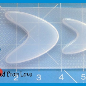 May include: Clear silicone mold with two boomerang-shaped cavities. The mold is rectangular with a textured surface and a small heart-shaped cutout. The text "Molded From Love" is visible in the lower left corner.