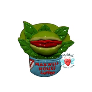 Pode incluir: Uma planta verde e vermelha com uma boca e dentes em uma xícara azul com o texto "Maxwell House Coffee" e as palavras "Molded from Love" em um coração rosa com asas.