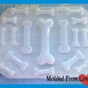 May include: A clear plastic mold with 13 bone shapes for making dog treats or other crafts. The mold is on a white cutting mat with a grid pattern. The text "Molded From Love" is visible in the lower right corner.