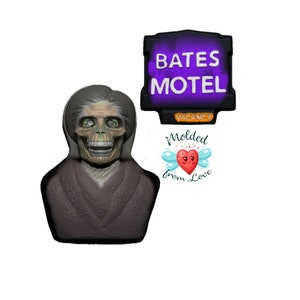 May include: A spooky Halloween-themed decoration featuring a skeletal figure and a sign that reads "Bates Motel". The sign has a purple background and the word "Vacancy" is visible. The figure is wearing a brown garment.