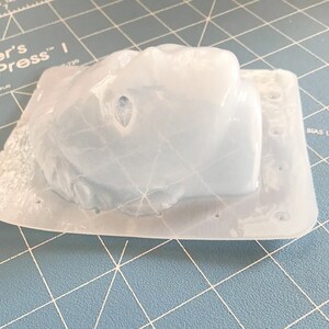 May include: A clear, molded plastic object, possibly a mold for crafting, sits on a blue cutting mat. The object has a rounded shape with a textured surface and is encased in a rectangular base. The plastic is translucent, allowing light to pass through.