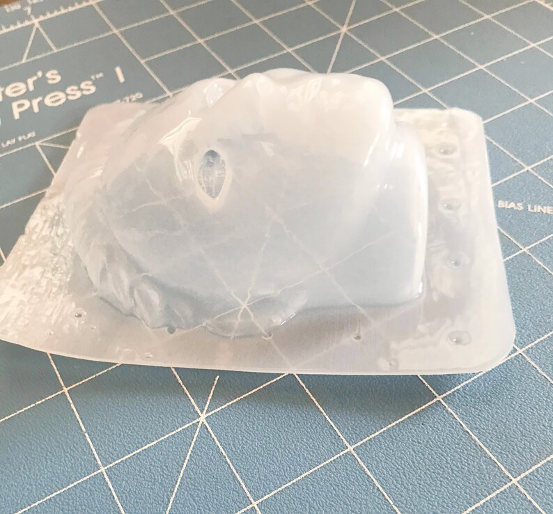 May include: A clear, molded plastic object, possibly a mold for crafting, sits on a blue cutting mat. The object has a rounded shape with a textured surface and is encased in a rectangular base. The plastic is translucent, allowing light to pass through.