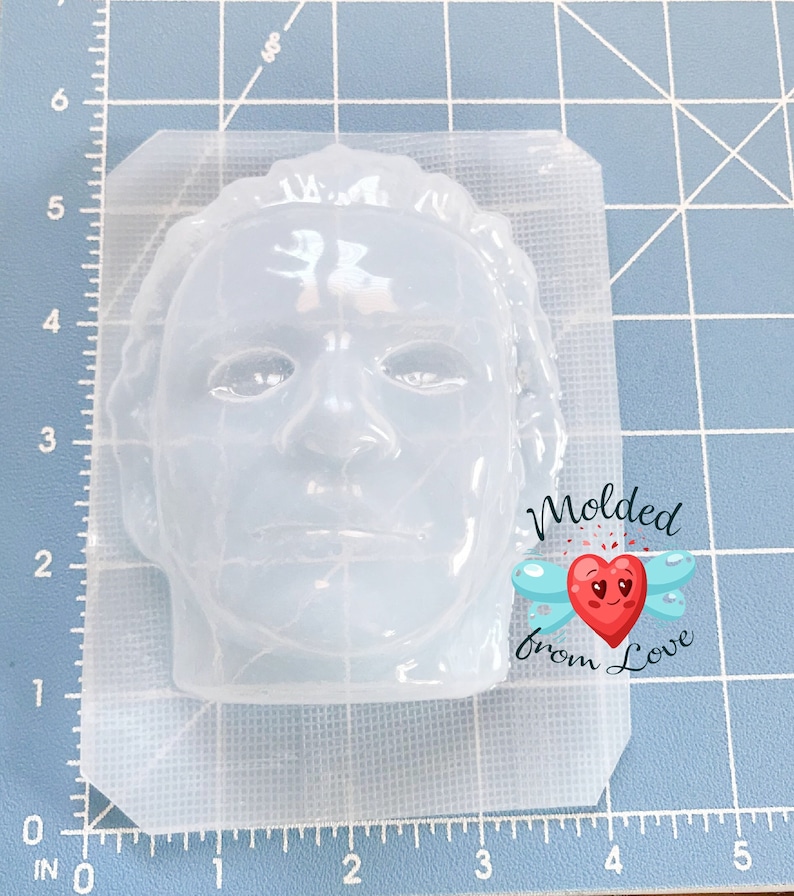 May include: A clear silicone mold in the shape of a face, with a detailed facial structure. The mold is on a blue grid surface. The text "Molded from Love" is on the right side of the mold.