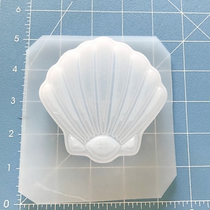 May include: A clear silicone mold shaped like a seashell. The mold is on a blue gridded surface.