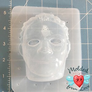 May include: A clear, flexible silicone mold in the shape of a face, likely for crafting. The face has detailed features, including eyes, a nose, and a mouth. The mold is rectangular with rounded corners. The text "Molded from Love" is visible.