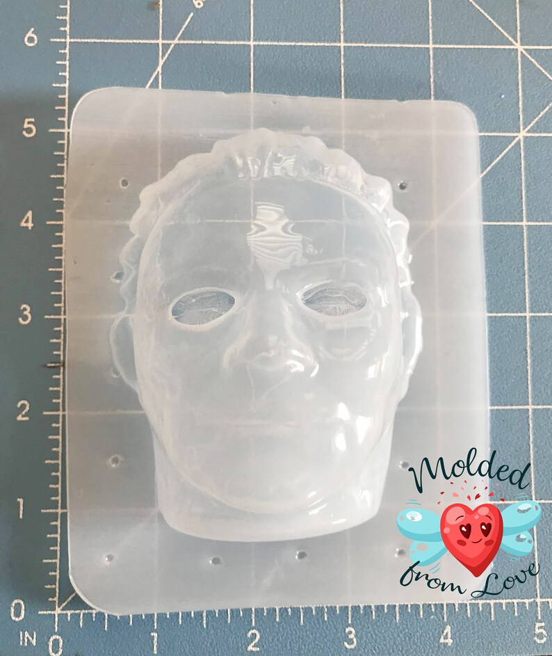 May include: A clear, flexible silicone mold in the shape of a face, likely for crafting. The face has detailed features, including eyes, a nose, and a mouth. The mold is rectangular with rounded corners. The text "Molded from Love" is visible.