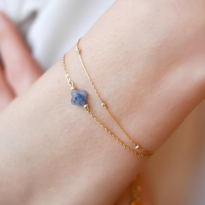 Puede incluir: Una delicada pulsera dorada de doble cadena. Una cadena presenta pequeñas cuentas redondas, mientras que la otra tiene un dije de flor azul de cuatro pétalos. La pulsera se lleva en la muñeca.